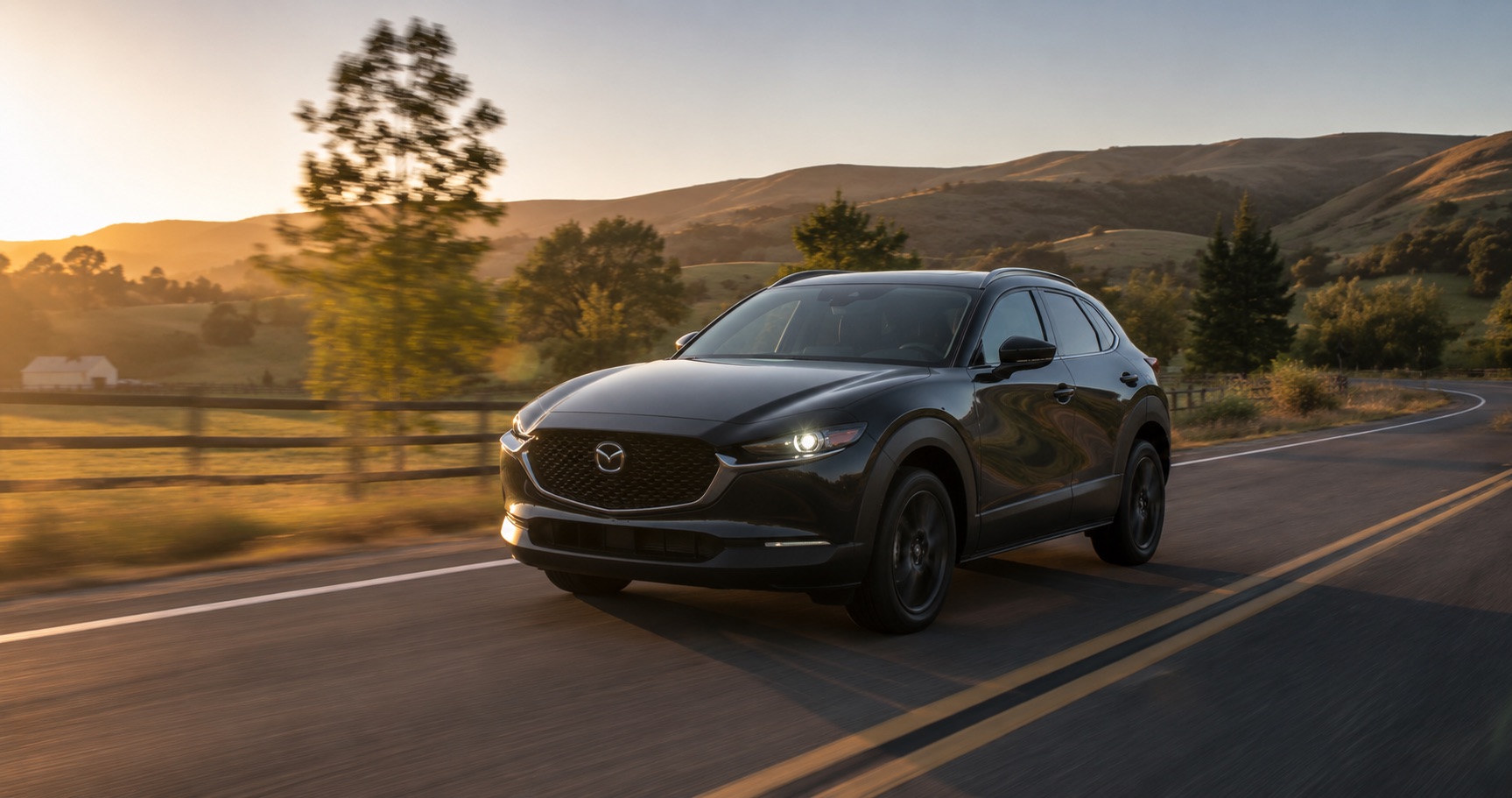 Black Mazda CX‑30 driving on winding country road at sunrise surrounded by hills and trees showcasing sleek front design and performance.