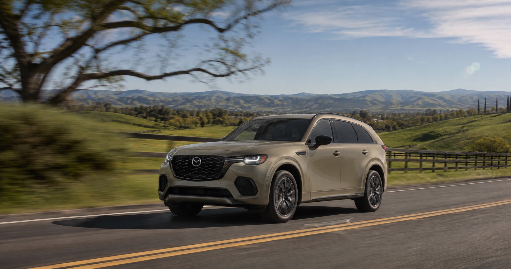 Tan-Green Mazda CX‑70 driving on winding country road surrounded by green hills and blue sky showcasing sleek design and performance.