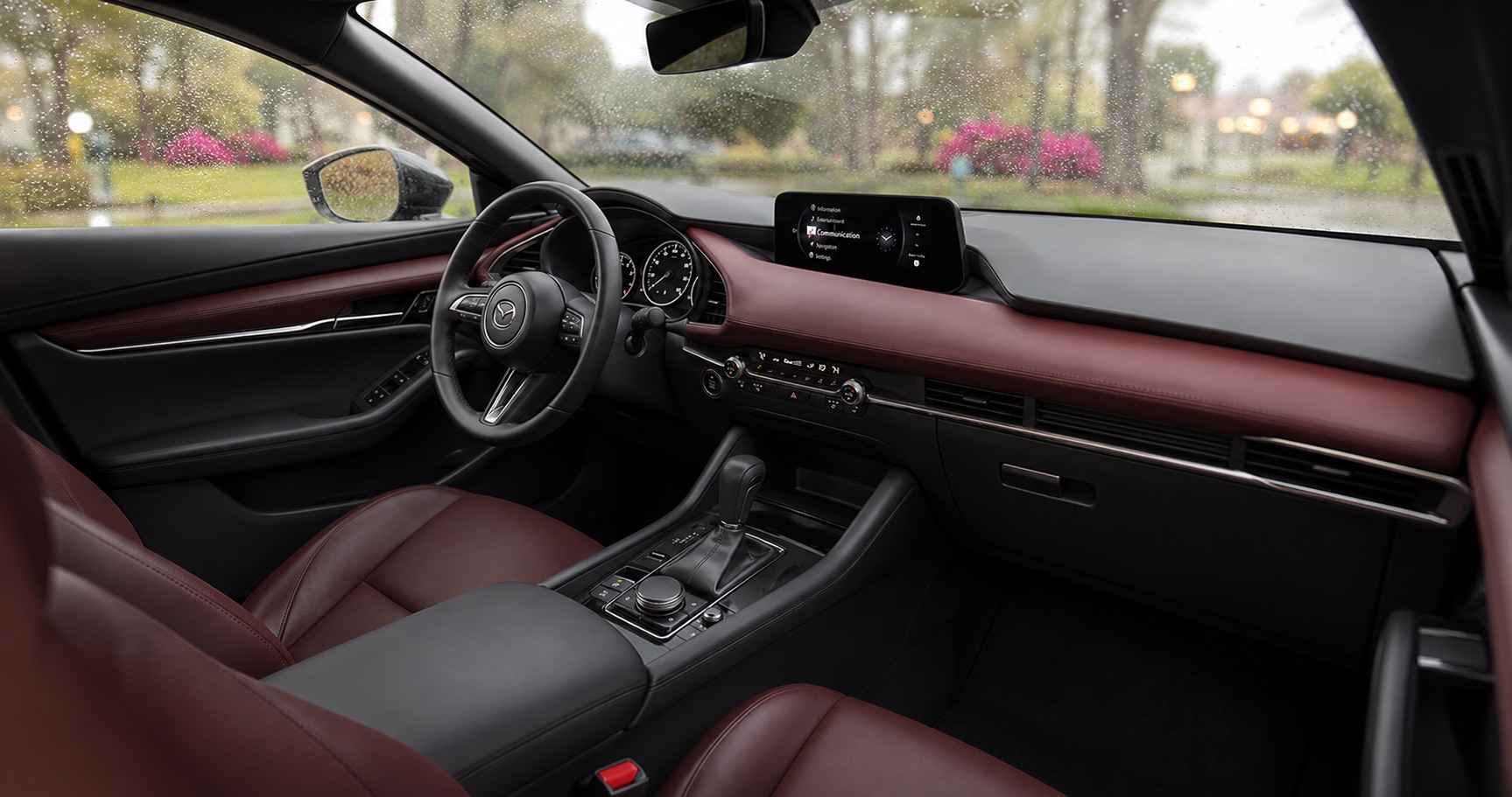 Mazda3 cabin with red leather seats and dashboard showing steering wheel center console and infotainment display with rain outside.