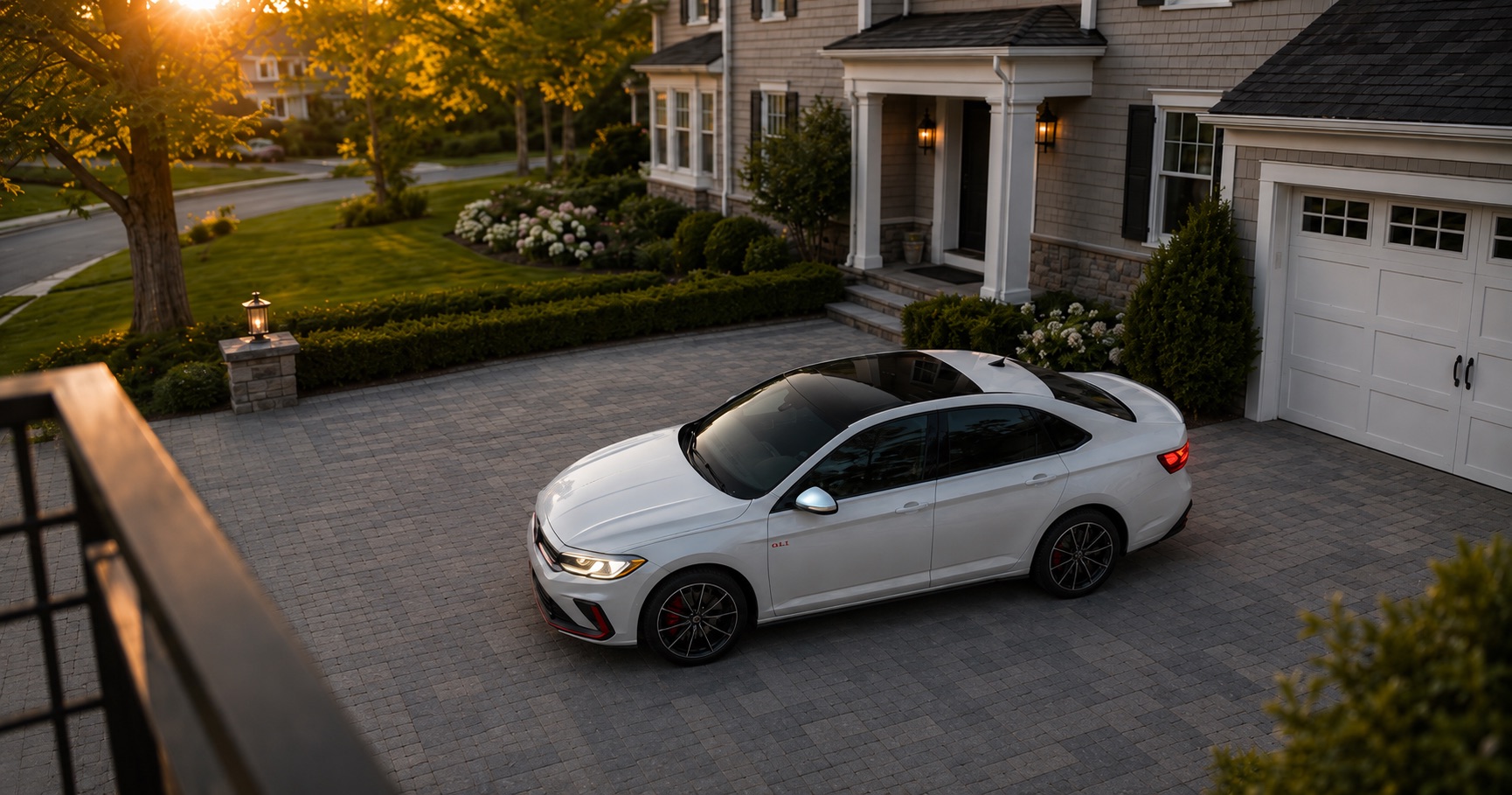 White Volkswagen Jetta GLI Autobahn parked in a driveway at sunset in front of a suburban home, highlighting its black roof, red brake calipers, and sporty design.