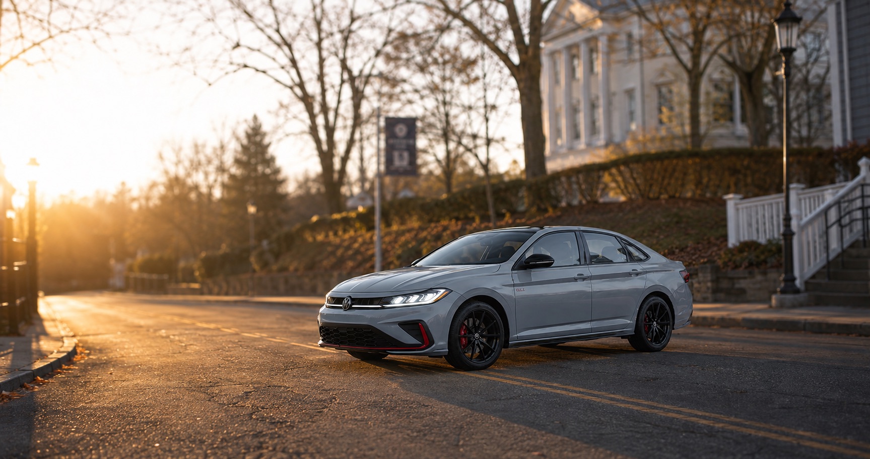 Gray Volkswagen Jetta GLI parked on quiet street near historic building at sunrise highlighting front grille red accents and premium trim details.