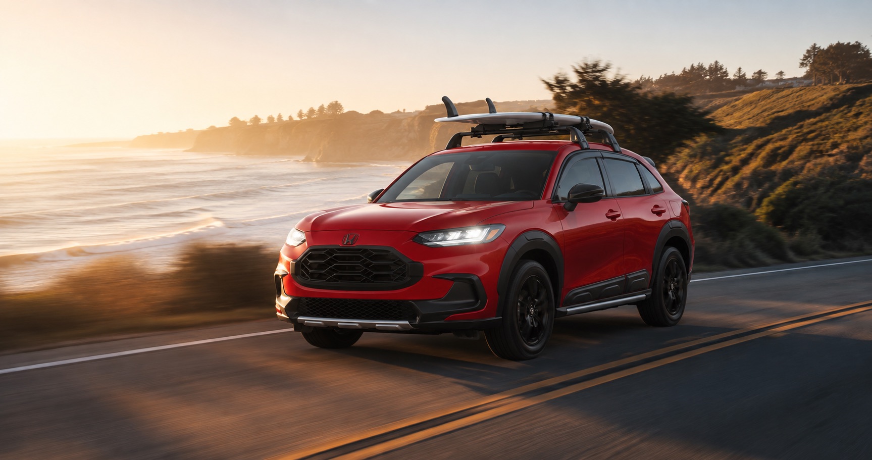 Red Honda HR-V driving along coastal highway at sunrise with surfboards mounted on roof emphasizing sporty styling and outdoor capability.