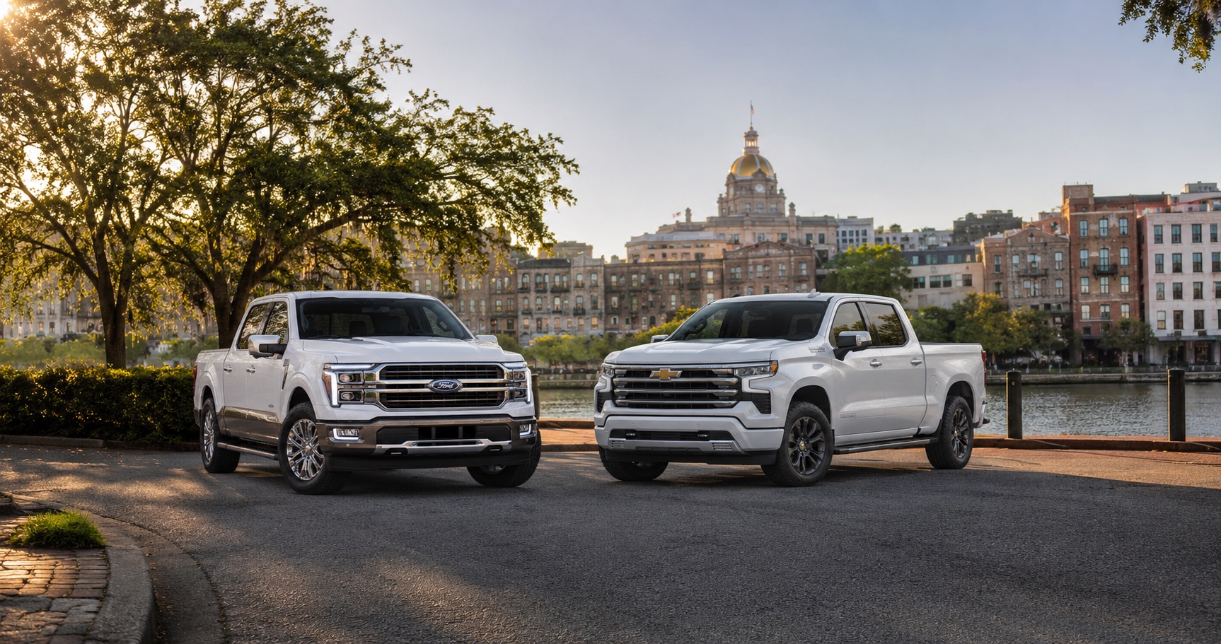 Two white pickup trucks parked side by side near the riverfront with historic buildings in the background.