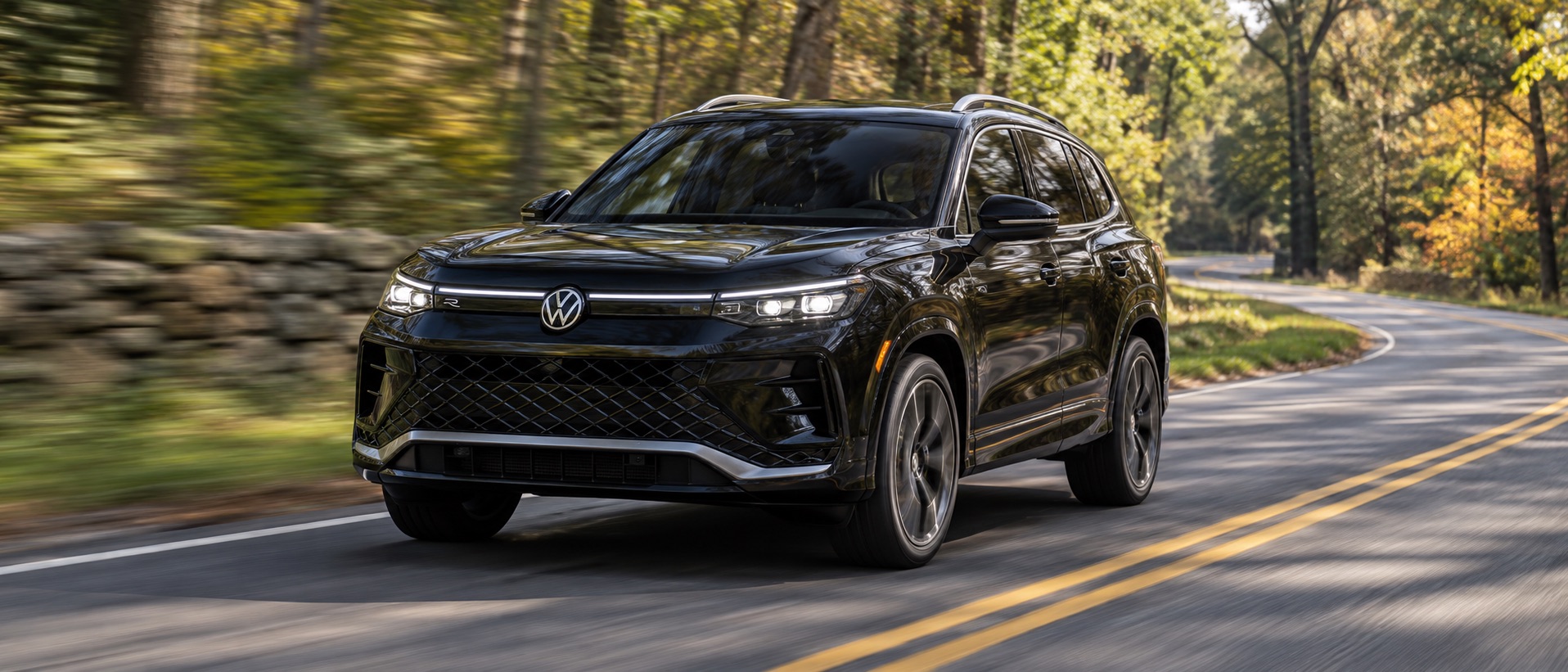 Black Volkswagen SUV driving on a winding tree-lined road with its LED headlights illuminated